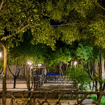 باغ در شب زیر سایبان درختان، با فانوس‌های روشن و نشیمن در انتهای مسیر